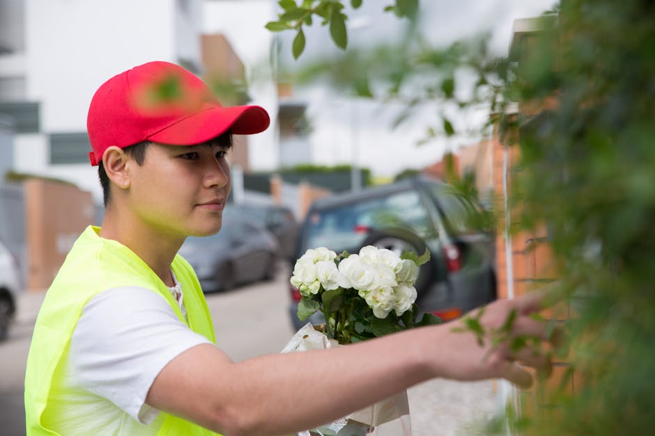 A young florist wearing a red cap and a fluorescent yellow vest is placing a fresh bouquet of white hydrangeas and roses against a wall in an outdoor setting, possibly preparing for flower delivery. T