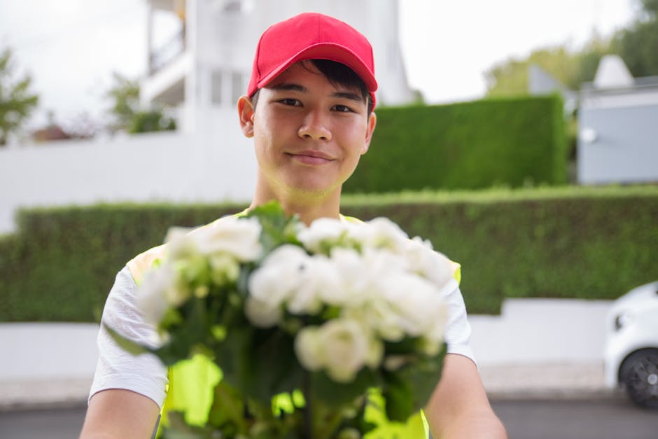 A delivery person wearing a bright yellow reflective vest and a red cap holds out a bouquet of fresh white roses and greenery towards the camera. The bouquet features tightly packed, vibrant white pet