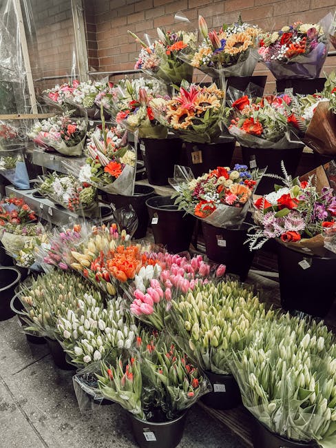 A variety of fresh flower bouquets displayed outdoors, featuring a colorful assortment of tulips, roses, lilies, and mixed blossoms arranged in black buckets and wrapped in black and transparent paper