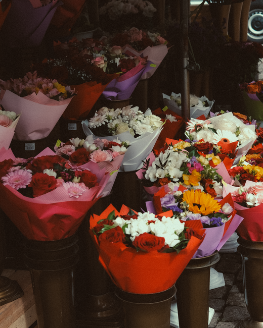A display of various floral bouquets arranged outdoors, featuring an assortment of fresh flowers including red roses, white lilies, pink carnations, yellow sunflowers, and purple statice. The bouquets