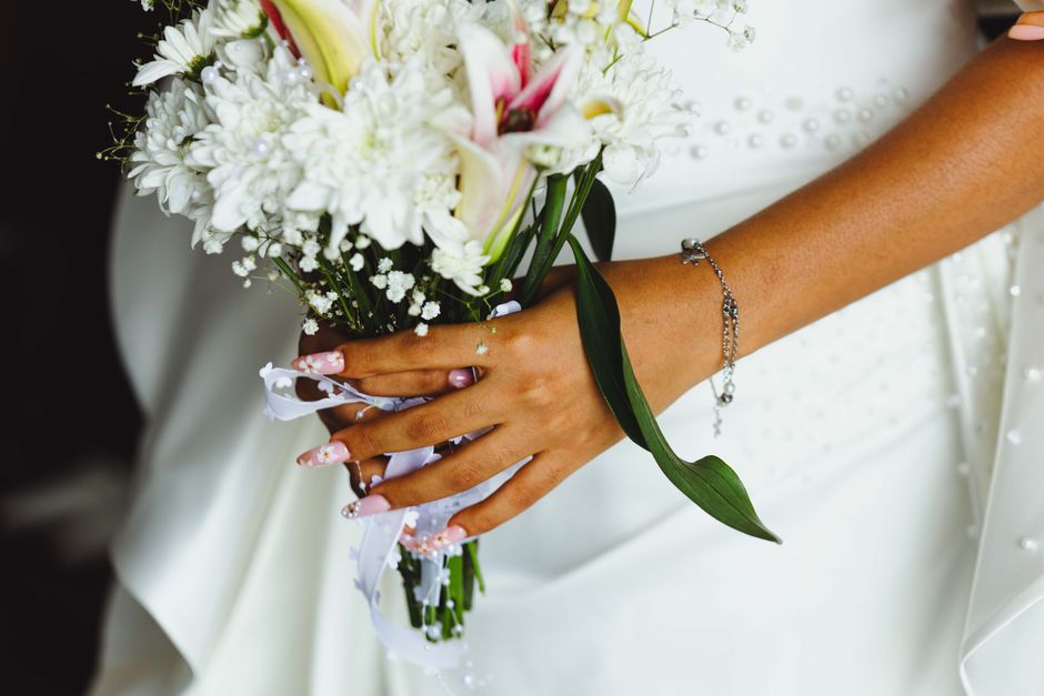 Close-up of a bride holding a bouquet of fresh white lilies, chrysanthemums, and baby's breath, wrapped in white paper with a satin ribbon. The bride's hand, adorned with a delicate silver bracelet an