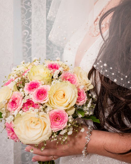 A bride holding a floral bouquet consisting of cream and pink roses, complemented by smaller white baby's breath flowers. The bouquet is arranged in a rounded style with a mix of open and partially op