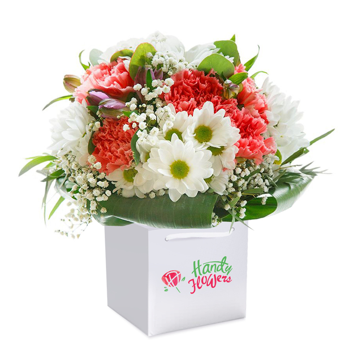 Capturing the spirit of Feltham's blooming parks, this impressive bouquet sits upright in a sleek white gift bag, its square shape a canvas for a riot of colour and freshness. At the centre, coral-pink and peach carnations unfurl in delicate, ruffled layers, their rich yet gentle hues reflecting the rosy glow of dusk seen from Bedfont Lakes. Enlivening the display, bright white daisies with distinctive lime-green centers stand out, their crisp petals forming a lively, textured contrast. Throughout the bouquet, fine sprays of white gypsophila add airy dimension and a sense of lightness, just like the flutter of petals in a summer breeze across Feltham. Emerging along the side, a few unopened purple-tinged buds echo the hope of new beginnings, infusing the arrangement with depth and a sense of natural progression. Framed by a generous collar of deep green foliage and broad, lush leaves, every flower shines against a backdrop that hints at local green spaces. The carefully balanced, rounded form speaks of expert hands and an understanding of harmonious composition. Presented in a chic white bag with strong ribbon handles, this bouquet is effortlessly giftable, making it the ideal choice for marking milestones, sending love, or brightening a home within Feltham's friendly neighbourhoods. The clean white background accentuates purity and vibrance, ensuring the arrangement's artistry and meaning come to the fore for local flower lovers.