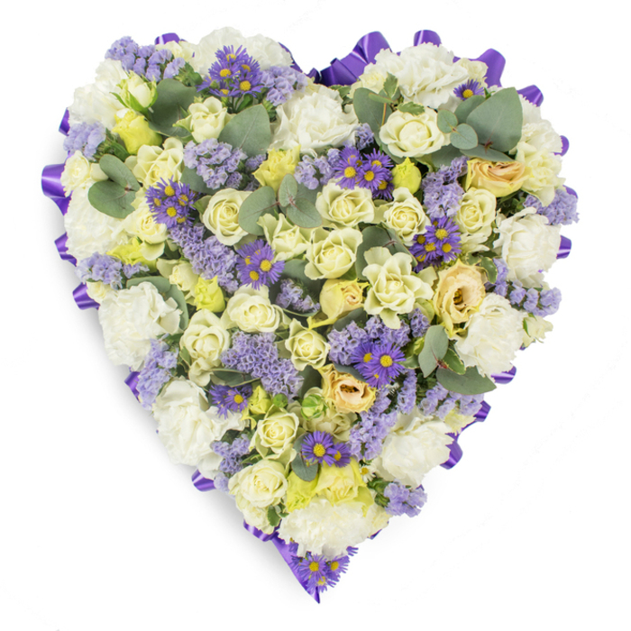 Presented on a spotless white backdrop, this heart-shaped floral tribute is a study in serene contrast and careful composition, designed to convey peaceful remembrance. A continuous ribbon of deep purple satin is looped and turned along the exterior edge to give the shape a poised, finished border. Within that frame, a soft field of white carnations provides generous volume and a cloud-like texture beneath plentiful cream and pale yellow roses-buds and gently opening blossoms with the slightest green-tinged tips that suggest freshness and gentle movement. Interwoven among the lighter blooms are clusters of subtle lavender statice and vivid small purple daisy-like flowers with bright yellow centers, which knit through the piece as visual punctuation and textural counterpoint. Rounded eucalyptus leaves in a cool blue-green are strategically placed to break the floral mass and introduce a modern, silvery accent and implied fragrance. The overall palette-whites, creams, pale yellows, and layered lavenders edged in richer purple-reads as compassionate and restrained, an appropriate and graceful choice for funeral services, wakes, or graveside memorials. Crafted with the care of an experienced local florist, this design would sit respectfully at services held near Hanworth Park or on Feltham High Street, offering families a clear, dignified expression of enduring love and heartfelt condolence.