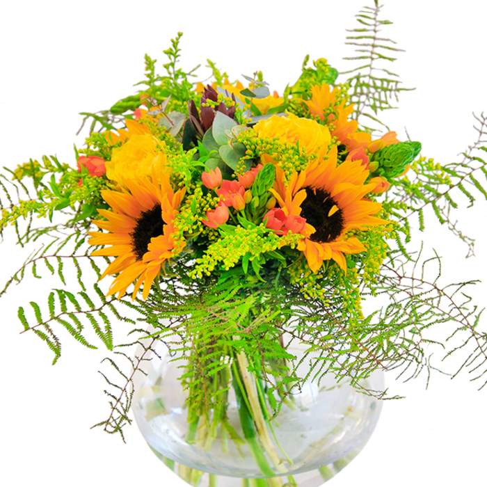 A dazzling fusion of sunshine yellow and autumn red, this bouquet makes an unforgettable entrance-its exuberant sunflowers and velvety yellow roses capturing the essence of a golden afternoon in Feltham. The sunflowers spread wide, petals bursting with warmth around deep mocha centres, while garden roses, packed in sumptuous layers, provide both classic structure and a dreamy softness. Ruby-red Hypericum berries are tucked throughout, shining among the foliage much like the vibrant berries that dot neighbourhood hedgerows. Cascading fronds of fern, light green filler, and whimsical eucalyptus leaves spill elegantly over the sides, their different shades and textures reminiscent of Feltham's riverside pathways and hidden green corners. All are set within a perfectly clear, round glass vase, its form echoing the gentle curves of water and sky over nearby parks. Crisp, fresh stems are on display beneath the water, further accentuated as natural light kisses the blossoms and leaves. The bouquet emits an understated, sweet fragrance-a whiff of luxury and nature combined. Meticulously arranged by Feltham Flowers, it's a fitting choice for moments of celebration, from birthdays to new beginnings, or simply sharing the spirit of West London's blooming landscapes with family and friends.