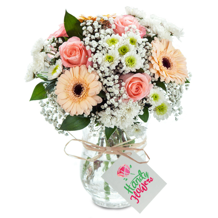 Capturing the inviting spirit of Feltham, this elegantly arranged mixed bouquet is proudly presented in a clear glass vase, its form defined sharply against a luminous white backdrop. At the centre, pastel peach-pink garden roses bloom with soft vitality, their spiral petals full of romance and classic appeal. These are framed by a pair of peach gerbera daisies whose bold forms and dark, defined centres bring an assertive optimism. Throughout, clusters of petite, white chrysanthemums with radiant lime centres and generous sprays of cloud-like gypsophila lend volume and a sense of airiness, reminiscent of Feltham's spring flower shows. Strategic leafy accents in rich green add depth, balancing the composition and highlighting the local florist's deft touch. Every stem is showcased through the vase's clear glass, their crisp lines visible in cool water-a subtle reminder of Feltham Flowers' dedication to freshness. Around the neck, rustic twine holds a plain white tag-ready for your message. This bouquet is equally at home as a housewarming gift, anniversary centrepiece, or heartfelt thank you, offering a gentle embrace of colour and community in every petal.
