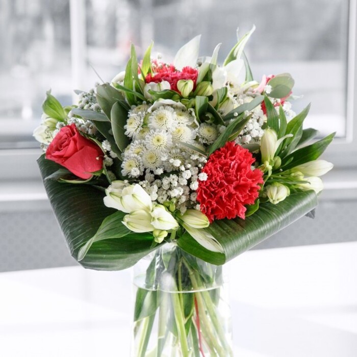 A graceful, modern bouquet in passionate red and tranquil white stands in a clear cylindrical vase, its stems crisply aligned and visible through the water like a living sculpture. At the heart of the arrangement are deep, velvety red roses, their petals just beginning to open, paired with a richly frilled crimson carnation that provides a focal point of intricate texture. Around these, white blooms offer a gentle counterpoint: alstroemeria with slim, speckled petals, soft chrysanthemum-style flowers, and closed white lily buds that promise to unfurl in the coming days, extending the life and interest of the design. Tiny clusters of gypsophila - baby's breath - float among the flowers, giving the impression of light mist or scattered stars, and lending the piece an ethereal, celebratory quality. The bouquet is carefully cradled at its base by broad, dark green leaves, polished to a glossy sheen and wrapped into elegant curves that create a defined collar and a contemporary edge. Behind and between the main flowers, assorted green foliage in eucalyptus-like tones and pointed shapes adds depth, structure, and an almost woodland freshness. Resting on a smooth white surface, with a softly blurred, bright background reminiscent of natural daylight filtering through a window in a Feltham living room near Bedfont Lakes Country Park, the arrangement feels both uplifting and serene. As a florist, the harmony between the bold reds and the quiet whites, and the way each stem supports the next, makes this a perfect centrepiece for sharing gratitude, love, or friendship - whether it's placed on a dining table for a family celebration or delivered across Feltham to mark an anniversary or heartfelt thank-you.