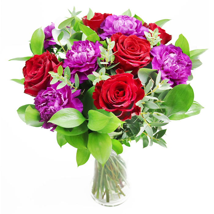 In the photograph, a luxurious mix of deep red roses and opulent purple peonies rises from a clear glass vase, the overall effect both classic and modern, much like a stylish Feltham townhouse balancing tradition with fresh energy. The roses are a saturated crimson, each head open to reveal a spiralling pattern of petals that feel plush and almost velvety to the eye, capturing that unmistakable language of romance. The purple peonies, by contrast, appear softer and more expansive, their many layers of fine, ruffled petals forming generous, cushion-like blooms that introduce an air of softness and indulgence. Surrounding these focal flowers is an array of lively foliage, with leaves of various shapes and textures-some broad and glossy, others slimmer and more delicate-creating depth and a natural, garden-inspired frame. Eucalyptus threads through the arrangement, its elongated stems and round, silvery leaves rising lightly above the main body of the bouquet, adding airy vertical lines and a subtle, contemporary twist that prevents the design from feeling too formal. The clear glass vase supporting the bouquet is elegantly understated, its transparency revealing the carefully aligned stems within and the clean water that anchors them, offering a glimpse of the unseen craftsmanship behind the arrangement. Gentle ambient light, similar to the brightness of a calm afternoon in Feltham or near Bedfont Lakes, washes over the flowers, intensifying the reds and coolening the purples in shadow. You can almost imagine leaning in to catch the combined fragrance of rose, eucalyptus, and earthy greenery-a scent at once soothing and uplifting. This bouquet would make a remarkable gift for an anniversary, a romantic celebration, or a milestone birthday celebrated in Feltham, bringing colour, texture, and an emotional warmth to the space it inhabits.