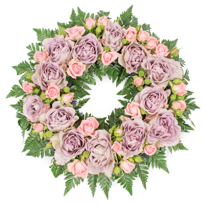 The image shows a beautifully composed sympathy wreath viewed from directly above, placed on a stark white background so that its form and colours are completely uncluttered. The design forms a flawless ring, with an empty centre encircled by a continuous band of flowers and foliage. The most prominent elements are large, fully opened roses in a refined mix of dusty lavender, soft mauve, and cool, grey-purple, each bloom revealing layers upon layers of gently curling petals. They are carefully spaced around the wreath so the eye can move evenly from one to the next. Between these focal roses sit clusters of smaller blush pink spray roses, their petals tighter and slightly more playful, offering a gentle contrast that softens the mood without losing the air of solemn respect. Here and there, small dark buds or fine, shadowy greens can be glimpsed between the flowers, giving the arrangement depth and a sense of natural growth. All the blooms rest on a thick base of dark green fern fronds, whose slender, serrated leaves fan outwards and inwards, framing the roses with feathery texture. The rich green tones anchor the soft purples and pinks, much like the mature trees and green spaces around Feltham give structure to the town's quieter streets. Soft, even lighting reveals the delicate gradients of colour and the freshness of the petals, suggesting a wreath recently crafted for a same-day funeral flower delivery. The overall impression is peaceful, dignified, and reassuring, the kind of floral tribute chosen in Feltham to convey love, gratitude, and remembrance when words alone do not feel enough.