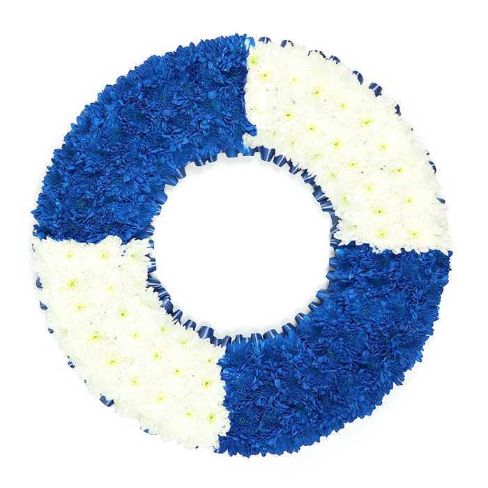 This image shows an elegant sympathy wreath crafted to resemble a classic lifebuoy, placed against a bright white background that makes its colours stand out vividly. The design is simple yet striking: a perfectly circular ring split into four equal sections that alternate between deep royal blue and crisp white flowers. The blooms, likely small chrysanthemums or similar compact varieties, are densely arranged so that the surface appears smooth and continuous, with a soft, cushion-like texture. The blue quadrants are rich and saturated, adding visual weight and symbolic strength, while the white sections bring brightness and a feeling of peace, much like clear daylight over nearby Feltham ponds or the open spaces of Hanworth Park. The ring is precisely shaped, with clean edges that reveal painstaking attention to detail by the florist. Around the inner circle, a slim band of dark blue ribbon is gently ruffled, tracing the curve and giving the impression of a real life ring without overshadowing the flowers themselves. The hollow centre remains uncluttered and perfectly round, acting as a calm focal point and subtly suggesting safe harbour or a final journey home. This floral lifebuoy is ideal as a funeral wreath or memorial tribute, especially meaningful for families honouring someone with links to the navy, lifeguarding, or maritime work, or a cherished relative who was simply a steady, supportive presence in their Feltham community. The balanced interplay of blue and white conveys messages of loyalty, guidance, and enduring love, turning this arrangement into a tender, symbolic expression of farewell.